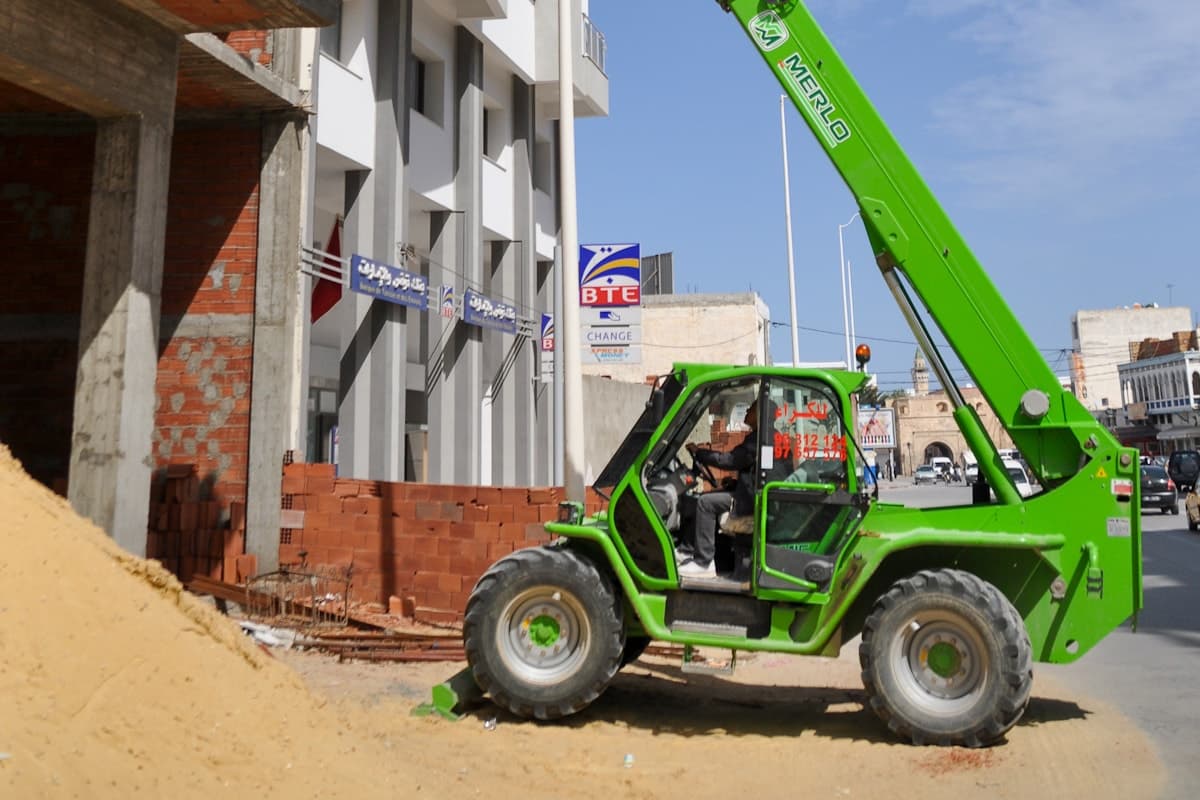 A counterbalance forklift on a warehouse floor next to a telehandler reaching over rough ground on a construction site